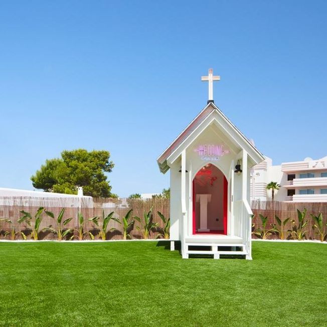 Capilla blanca sobre césped verde bajo un cielo azul, con una puerta roja y una cruz en la parte superior.