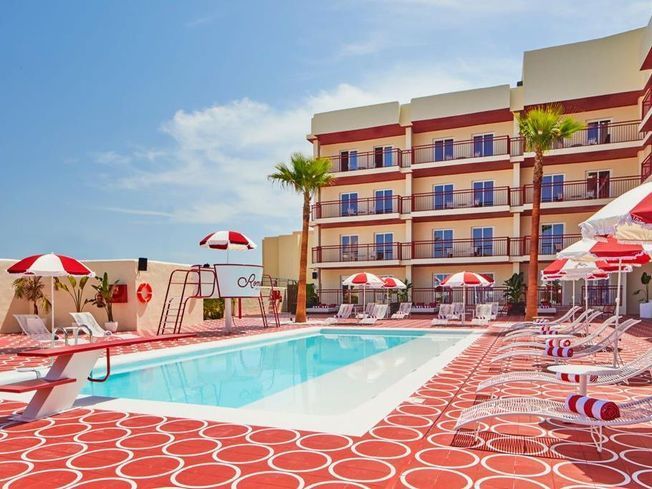 Vista de la piscina de un hotel con detalles en rojo y blanco, puesto de salvavidas, palmeras y tumbonas.