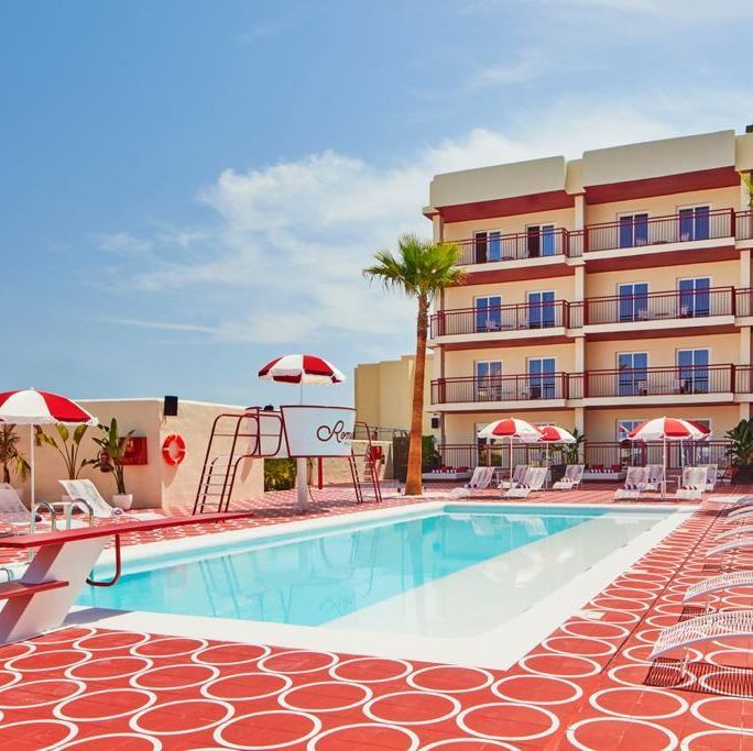 Escena junto a la piscina, sombrillas rojas y blancas y un edificio de hotel de color beige.