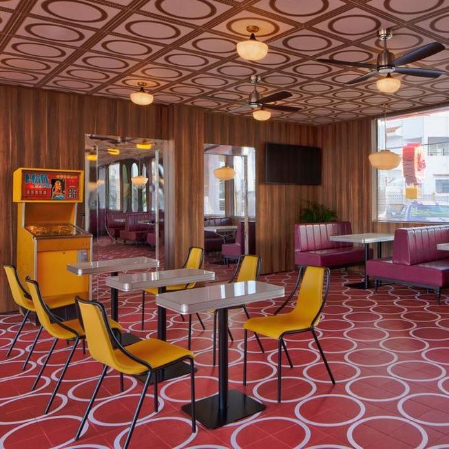 Interior de restaurante retro con tonos rojos, amarillos y madera. Mesas, sillas, cabinas, máquina recreativa y suelo y techo con motivos circulares.