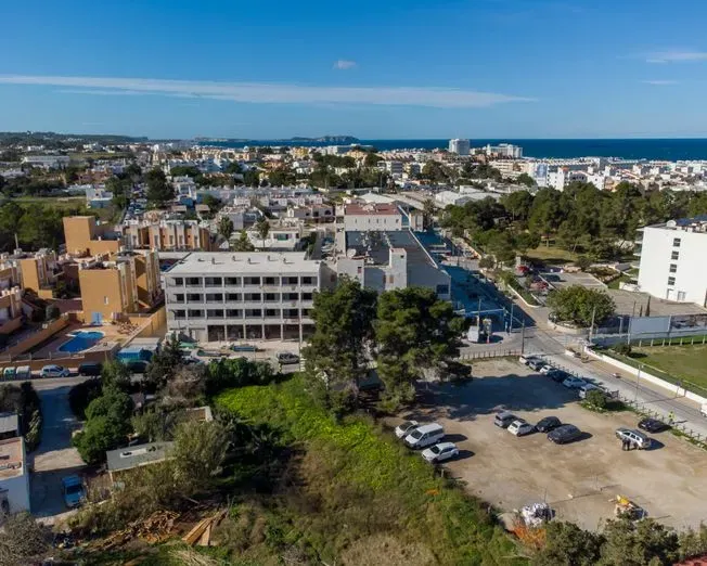 Vista aérea de una ciudad con edificios y un estacionamiento; cielo azul y mar al fondo.