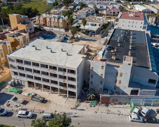 Edificio parcialmente construido en una zona urbana. Exterior blanco sin terminar, con techo oscuro. Coches aparcados cerca.