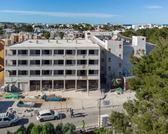 Construcción de un edificio de varios pisos bajo un cielo soleado; automóviles y trabajadores en la calle abajo.