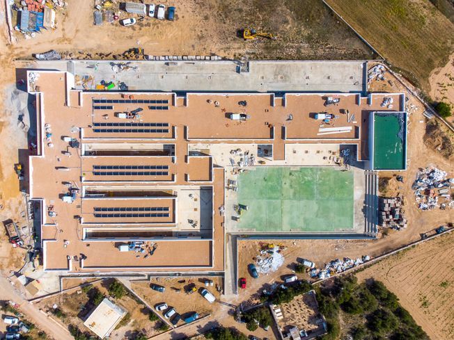 Vista aérea de un edificio en construcción con un campo verde y una piscina. Hay materiales de construcción dispersos.