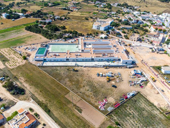 Vista aérea de una obra en construcción con un gran edificio y una piscina, rodeada de campos y un pequeño pueblo.