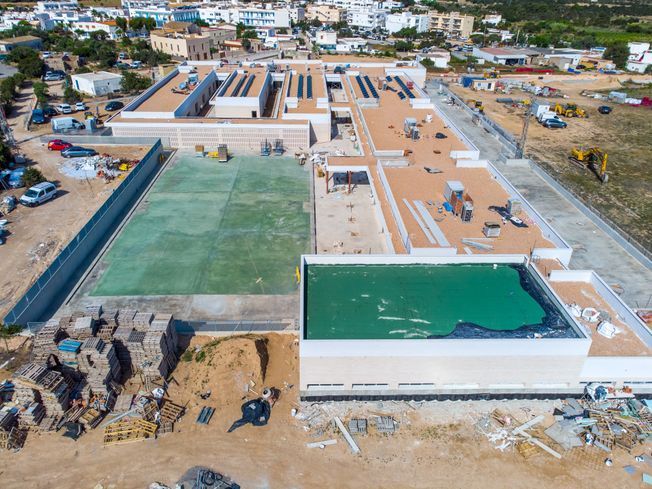 Vista aérea de un edificio en construcción con piscina y cancha de tenis; estructuras de color blanco y tostado.