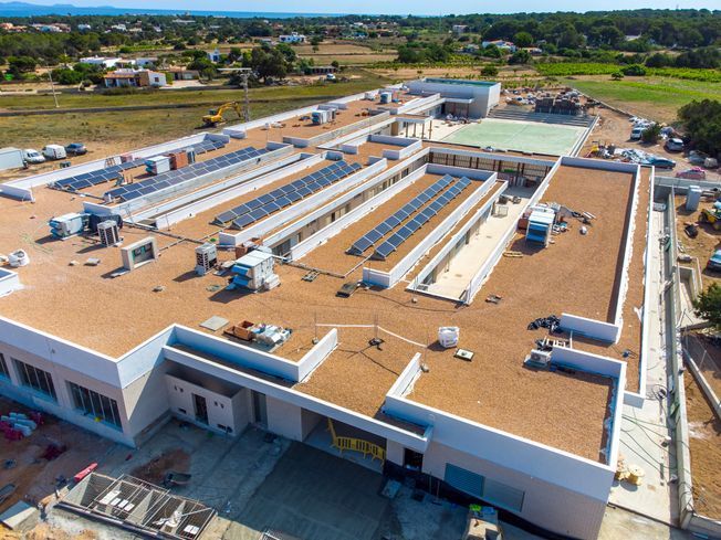 Vista aérea de un gran edificio en construcción, techo con paneles solares, rodeado de campo y otros edificios.