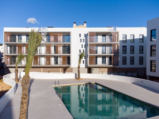 Complejo de apartamentos con piscina, exterior blanco, mamparas de madera en los balcones y palmeras.
