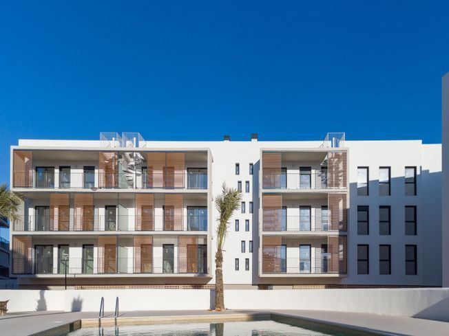 Edificio de apartamentos blanco con cortinas de madera, balcones y piscina en un día soleado.