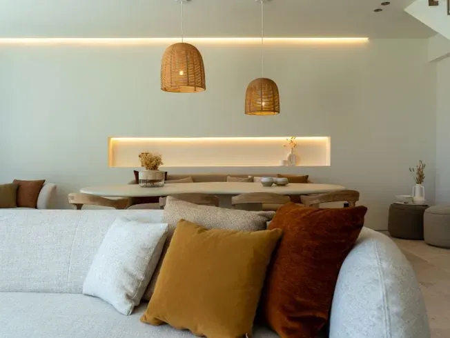 Sala de estar con colores neutros. Sofá con cojines decorativos, mesa de comedor de madera y lámparas colgantes tejidas.