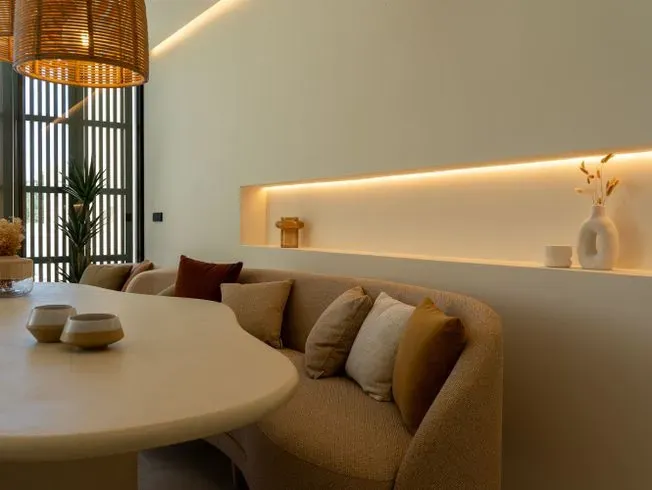 Acogedor rincón de comedor con un banco curvo beige, un estante incorporado con iluminación cálida y una mesa redonda.