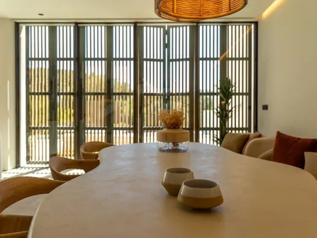 Comedor con mesa grande de color claro, sillas marrones y puertas-ventanas con listones.