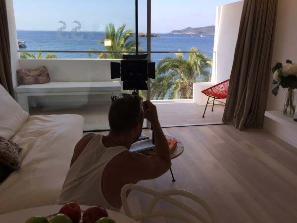 Hombre filmando con una cámara sobre un trípode en un interior con una vista panorámica del océano desde un balcón.