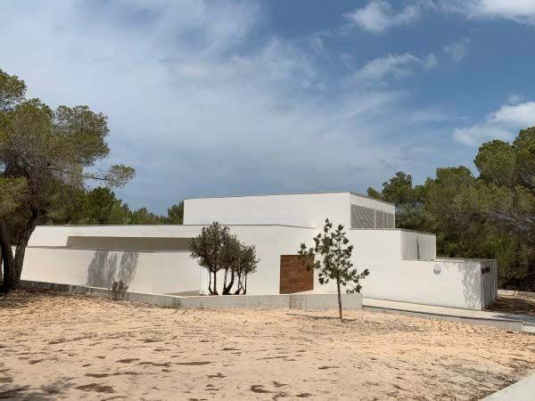Exterior de un edificio blanco moderno con una puerta de madera, un espacio de estacionamiento para discapacitados y árboles; el texto dice