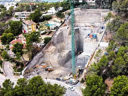 Obra en la ladera con una gran grúa, maquinaria y roca expuesta. Casas al fondo.