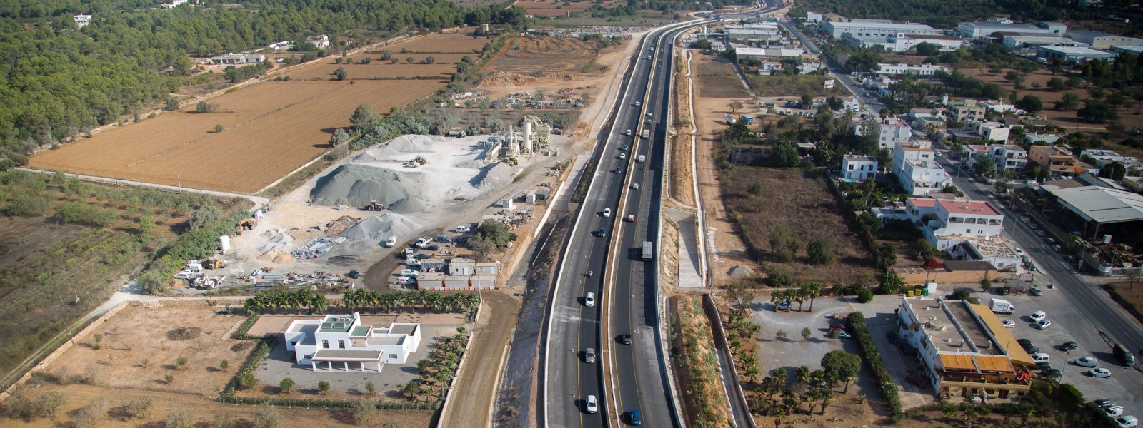 Vista aérea de una carretera de varios carriles que atraviesa un paisaje con edificios, árboles y campos secos.