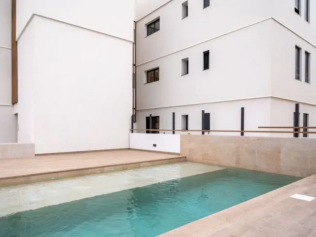 Una piscina de agua turquesa junto a un edificio blanco. La luz del sol proyecta sombras en las paredes.