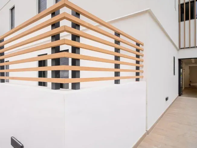 Exterior de edificio blanco con barandilla de color marrón claro, pilares y puerta de color negro.