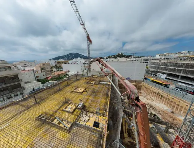 Obra: Se vierte hormigón sobre el tejado de un edificio con una grúa. Cielo nublado, paisaje costero.
