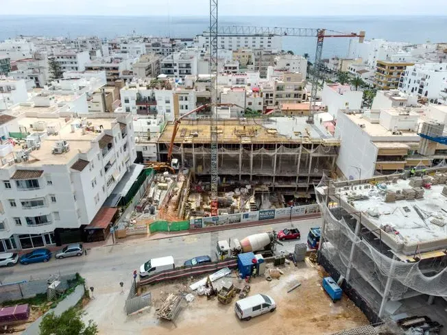Obra en construcción en una localidad costera con edificios, equipamientos y el mar visible al fondo.