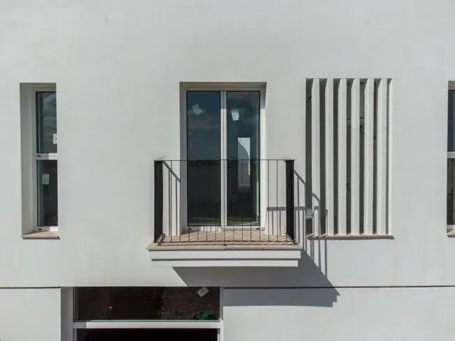 Fachada de edificio blanco con ventanas, balcón y lamas verticales que proyectan sombra.
