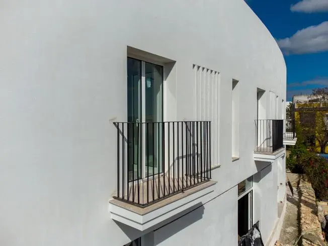 Edificio blanco con balcones, barandillas negras, puertas de cristal, cielo azul al fondo.