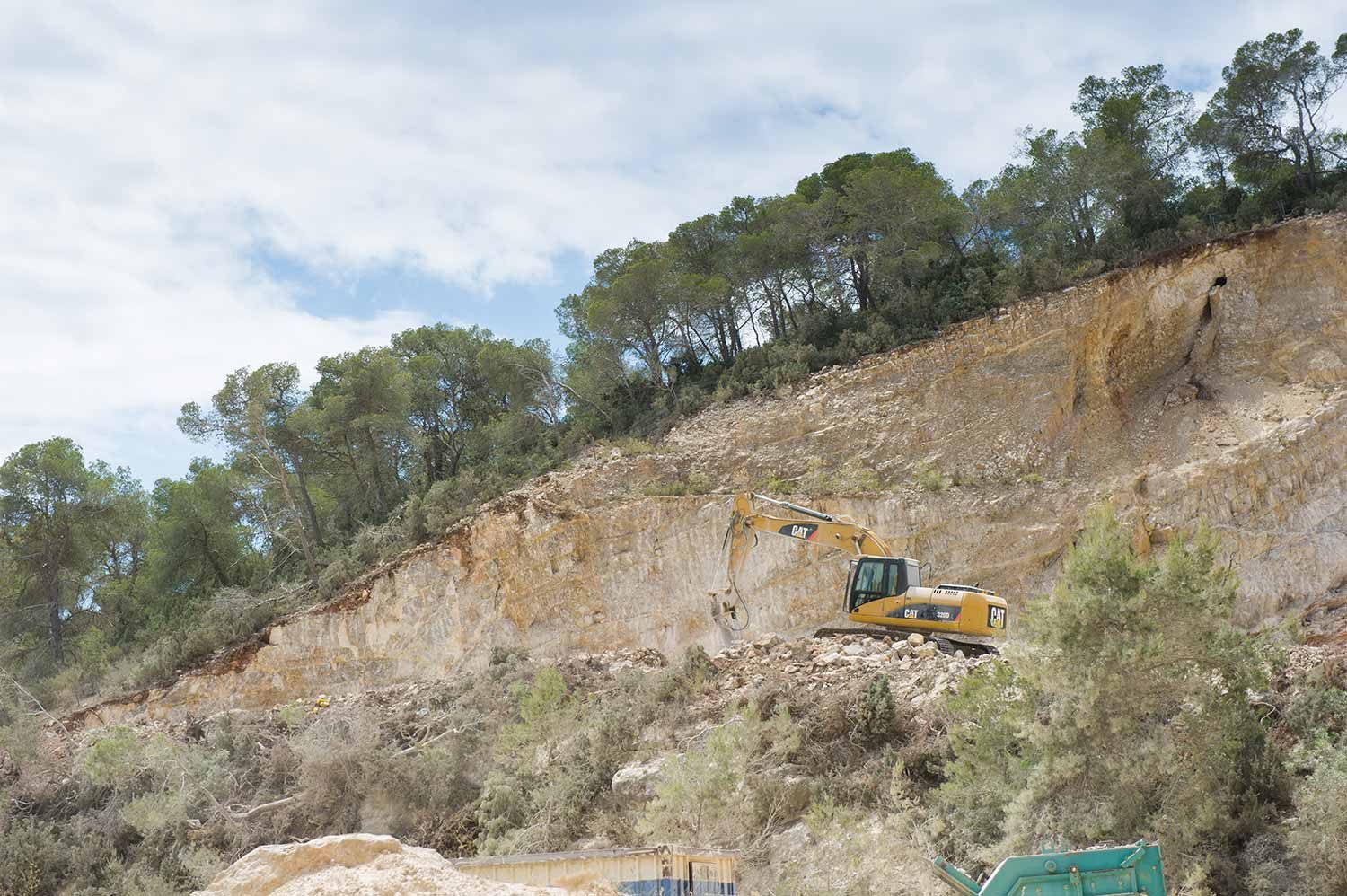 Excavadora en una ladera, quitando tierra de una pared de roca, árboles y cielo visibles.