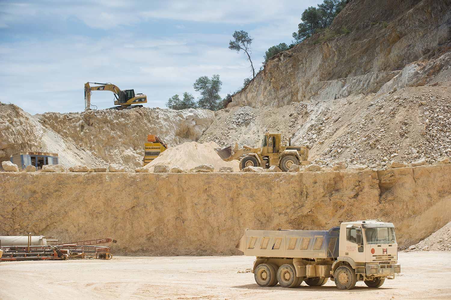 Escena de cantera con maquinaria pesada: excavadora, cargadora, camión volquete, trabajando en una pared de roca beige bajo un cielo azul.