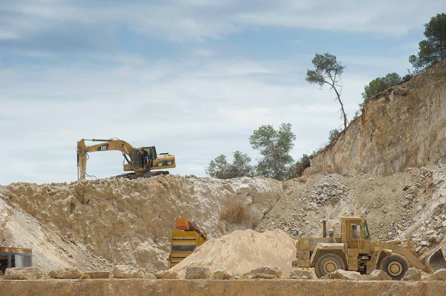 Excavadora y cargadora frontal trabajando en una cantera con suelo de color claro y un cielo parcialmente nublado.