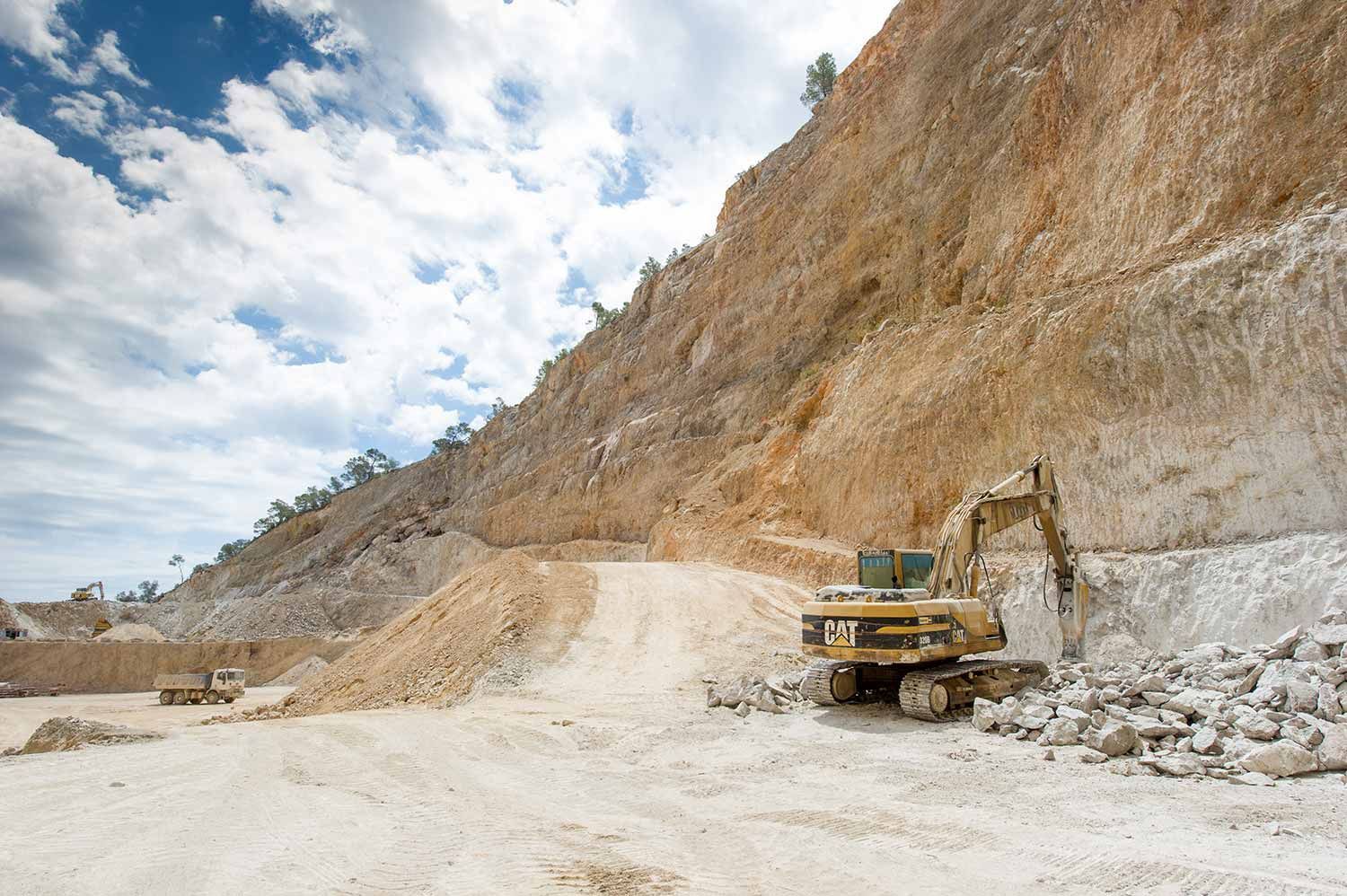 Excavadora en una cantera; una gran pared de roca con un camino y montones de rocas bajo un cielo nublado.