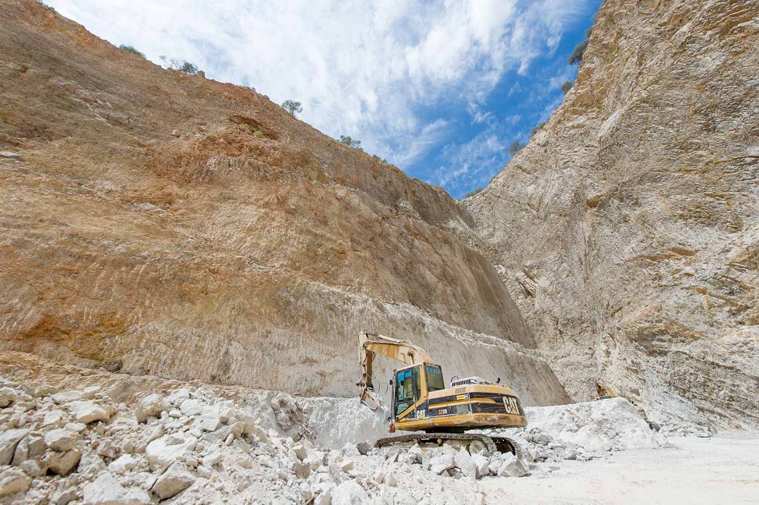 Excavadora extrayendo material en una cantera con acantilados de color beige bajo un cielo azul.