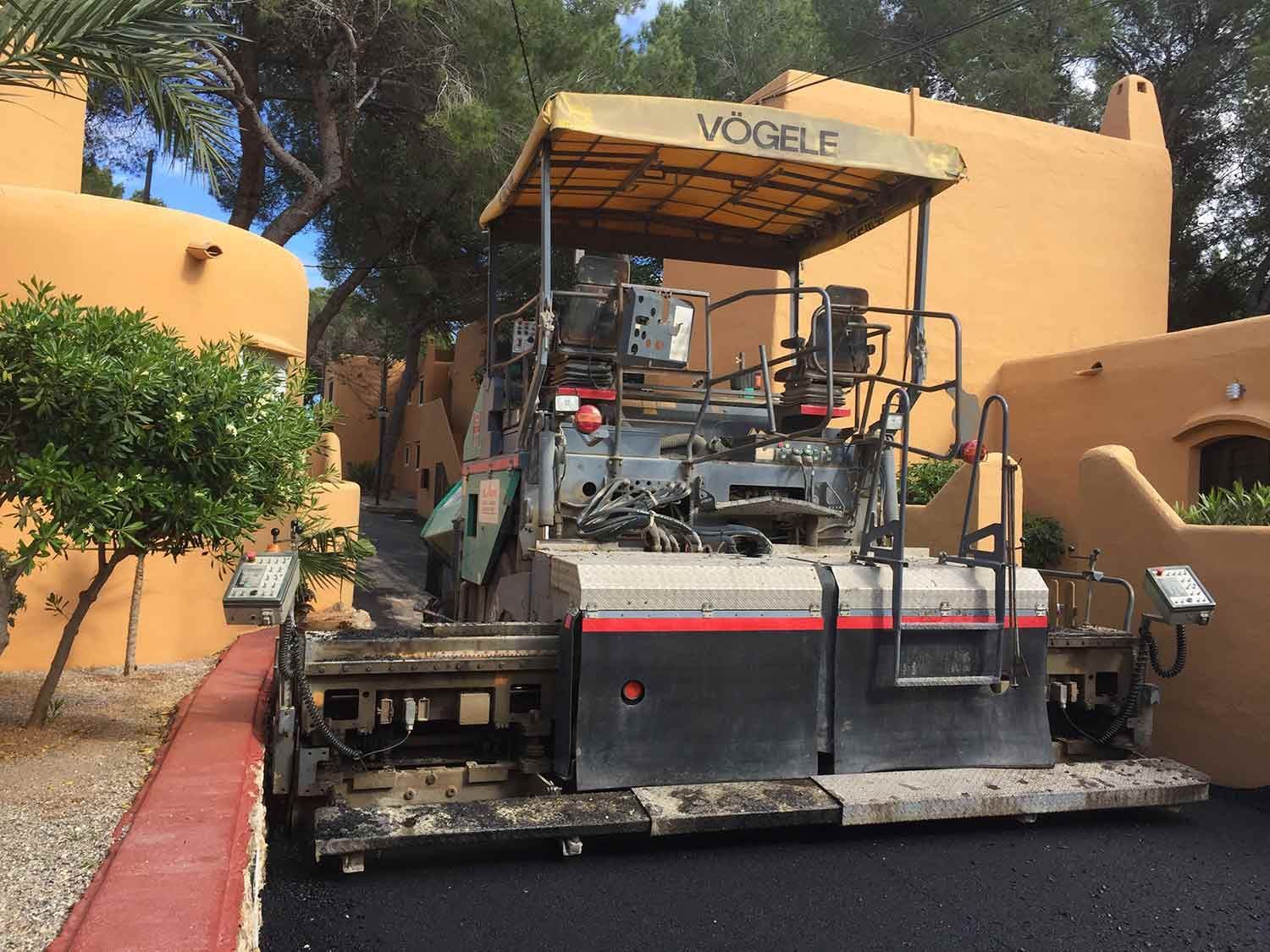 Pavimentadora de asfalto sobre una superficie recién pavimentada junto a un edificio de terracota. Día soleado.