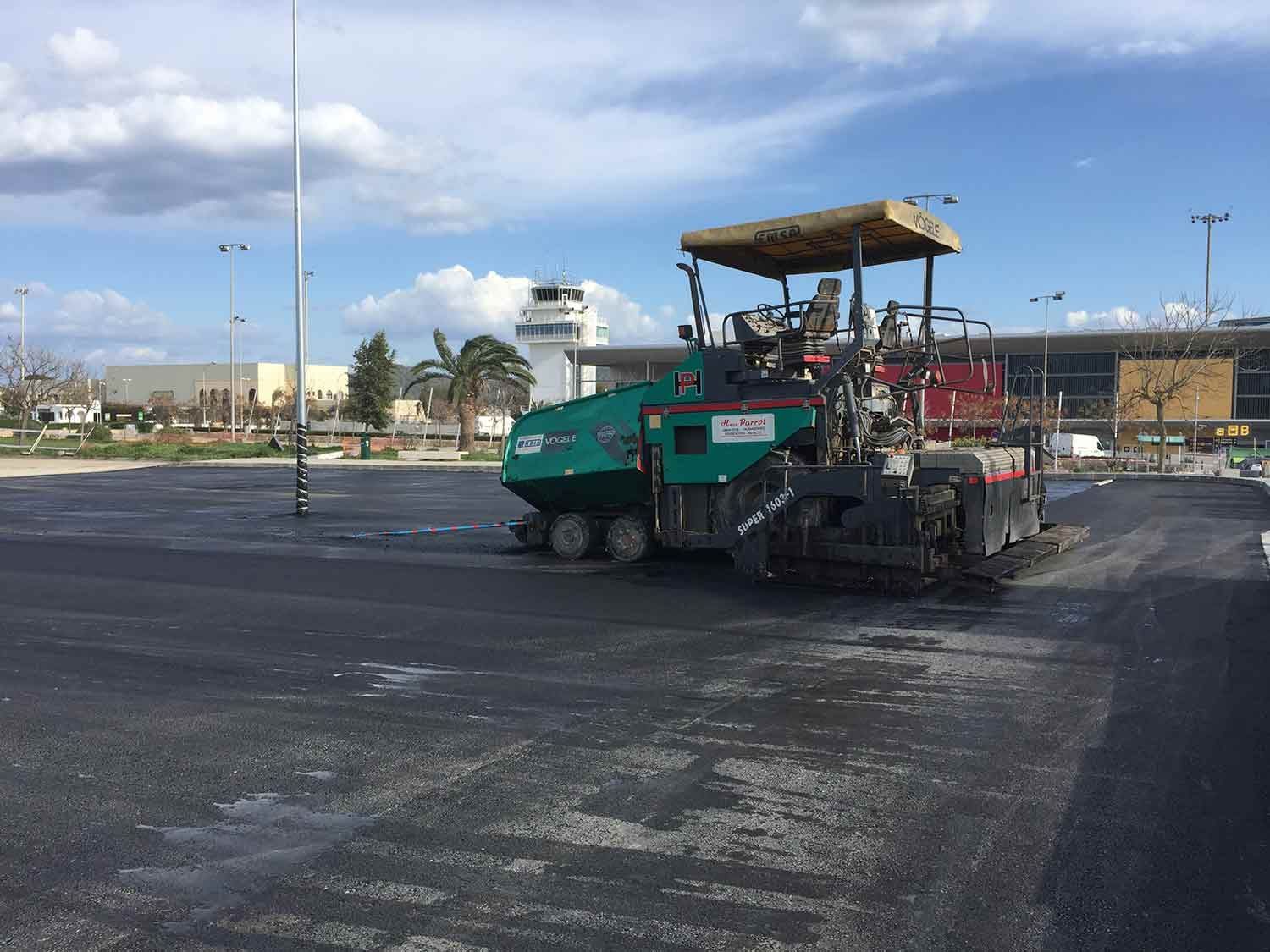 Máquina pavimentadora de asfalto colocando asfalto fresco en un área al aire libre, con un aeropuerto al fondo.