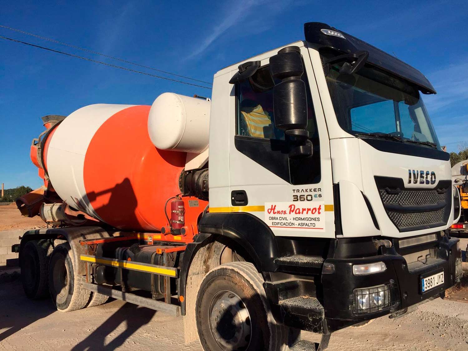 Camión hormigonera estacionado en un sitio de construcción, cabina blanca, tambor naranja y blanco.