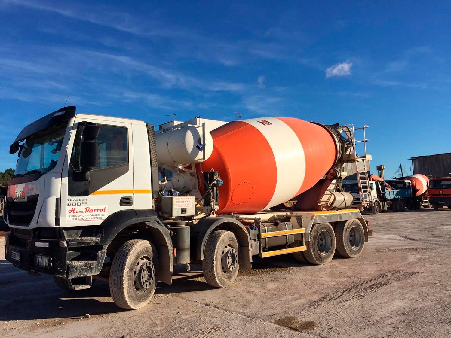 Camión hormigonera estacionado en un terreno de grava bajo un cielo azul. El tambor es blanco y naranja.