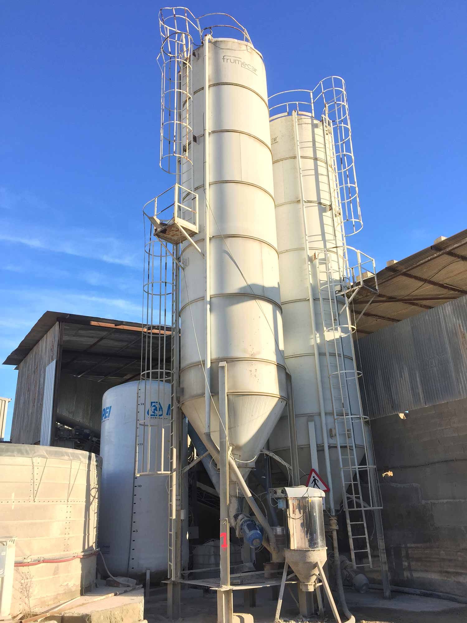 Afuera hay tres grandes silos cilíndricos con escaleras, al lado de un tanque más pequeño y un cielo azul.