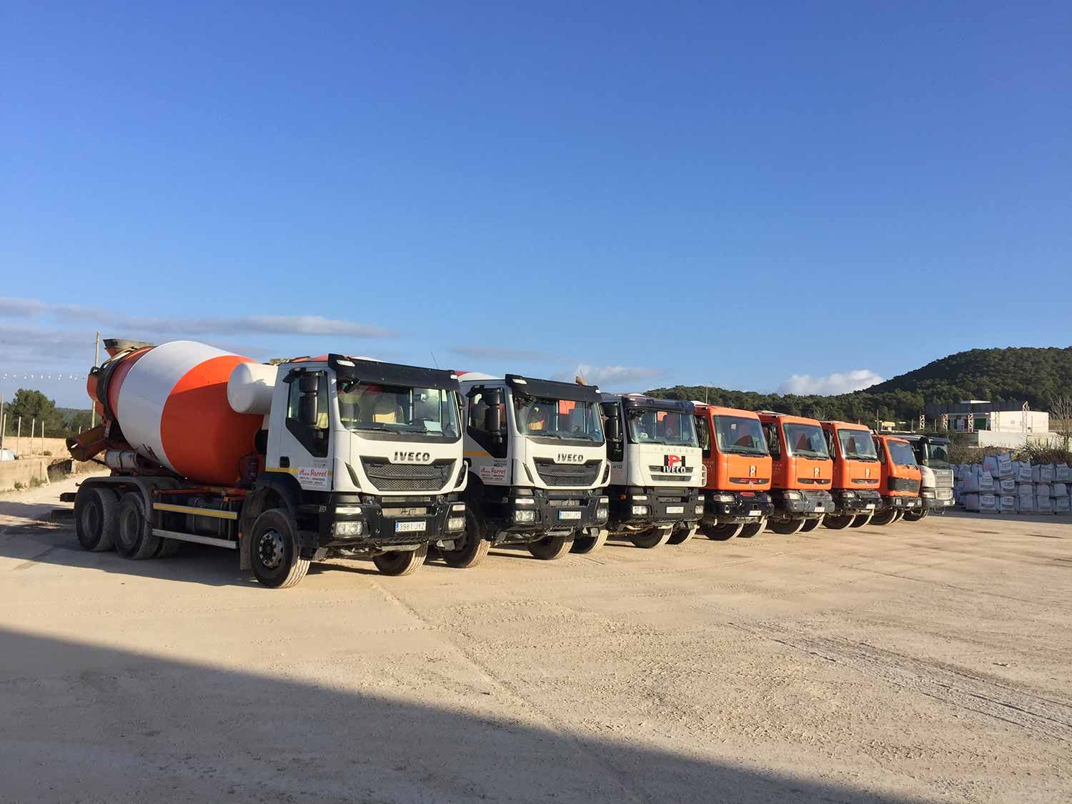 Una fila de camiones de cemento estacionados en un lote de grava bajo un cielo azul claro.