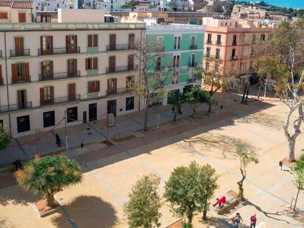 Plaza con edificios de varias plantas en la ciudad de Ibiza, España. Cuenta con árboles, bancos y peatones.