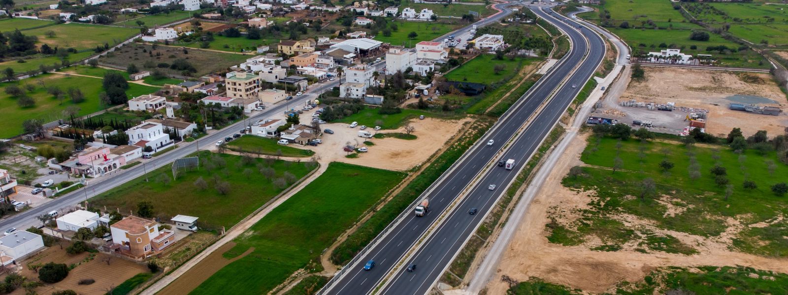 Vista aérea de una autopista que atraviesa un pueblo, con coches circulando. Campos verdes y casas bordean la carretera.