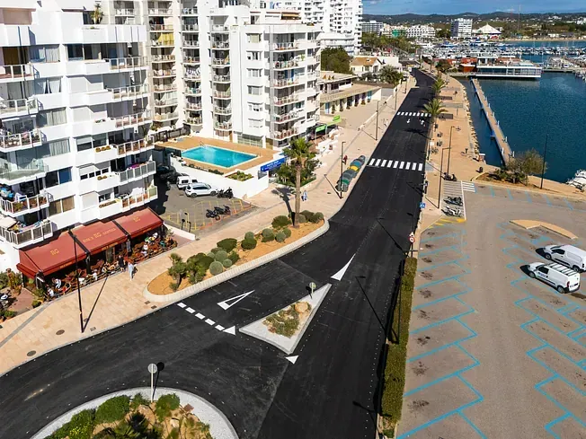 Vista aérea de una ciudad costera con puerto deportivo, carretera y rotonda. Edificios blancos, barcos y agua azul.