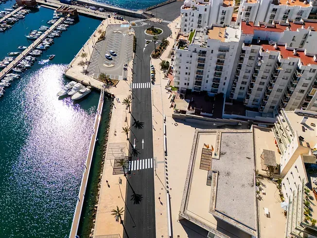 Vista aérea de un puerto deportivo con barcos, carretera y edificios en un entorno costero soleado.