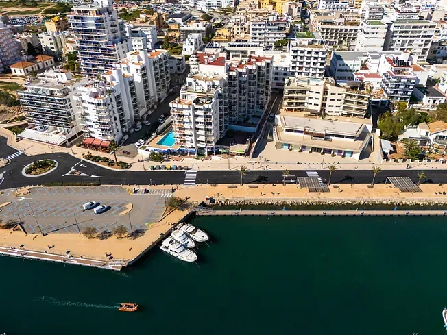 Ciudad costera, edificios, puerto con barcos, una rotonda y agua azul bajo un cielo soleado.