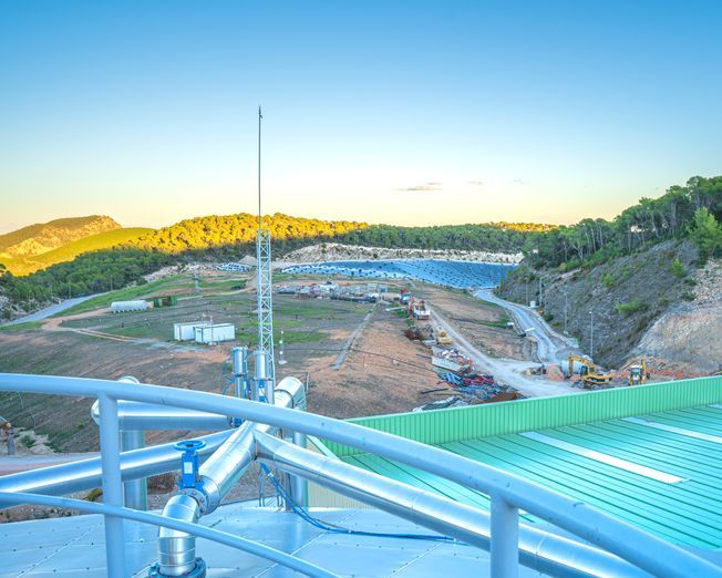 Instalación industrial con tanques, tuberías y un paisaje lejano con colinas, construcciones y paneles solares.