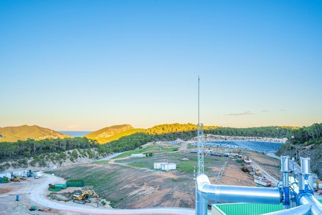 Vista aérea del sitio de construcción cerca de las montañas y el mar, con cielo azul y tuberías en primer plano.