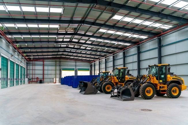 Dentro de un gran almacén, tres vehículos de construcción de color amarillo están estacionados sobre hormigón.