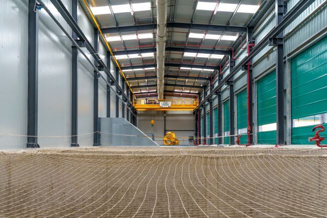 Vista interior de un gran edificio industrial con una grúa, paredes grises y una red de seguridad en primer plano.