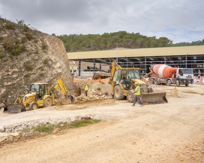 Sitio de construcción con retroexcavadoras y hormigonera, cerca de una ladera rocosa y un edificio.