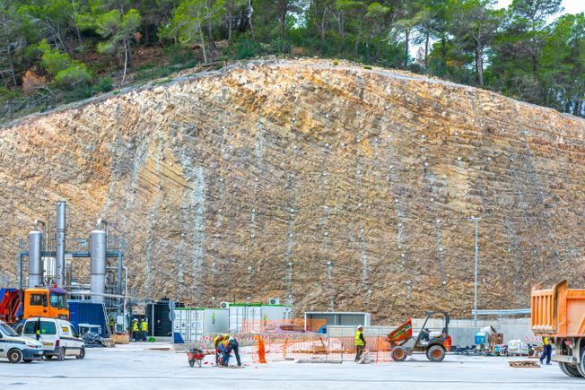Obra con trabajadores, pared rocosa y maquinaria. Árboles en la ladera.