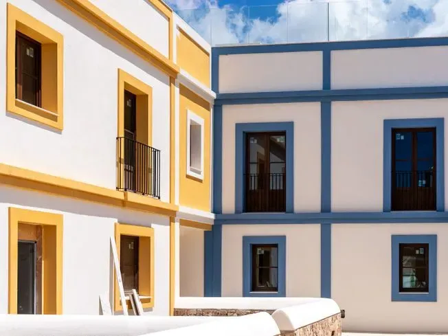 Edificio de dos pisos, mitad molduras amarillas, mitad molduras azules, paredes blancas, ventanas y cielo azul con nubes.