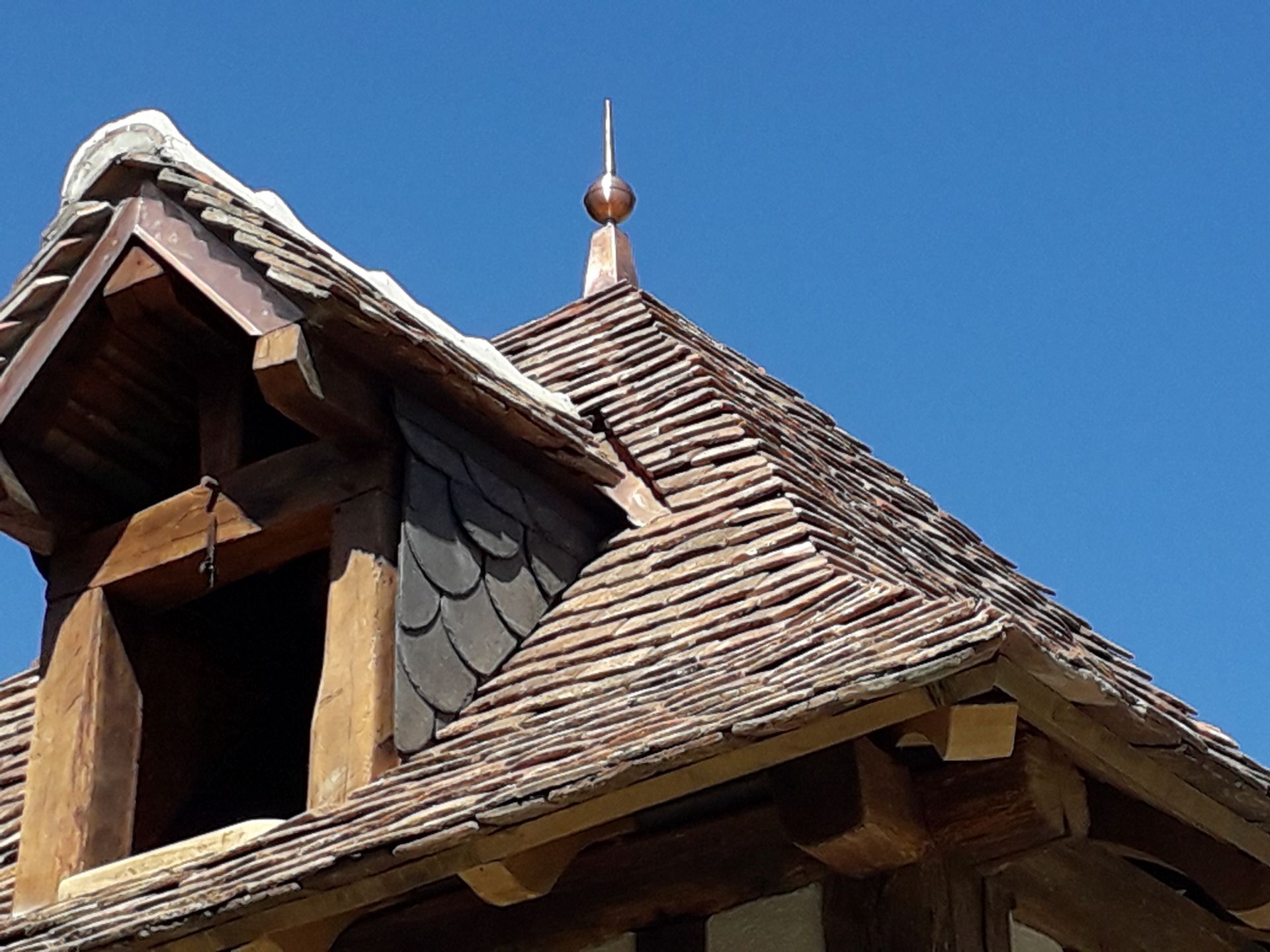 Toit en bois patiné par le temps, recouvert de bardeaux gris et surmonté d'une petite flèche, se détachant sur un ciel bleu.